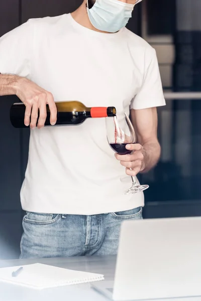 Vista recortada del hombre en la máscara médica verter vino en vidrio cerca de la computadora portátil en la encimera - foto de stock