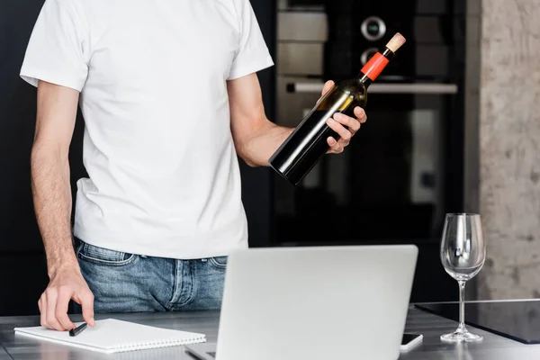 Vista recortada del hombre sosteniendo botella de vino cerca de la computadora portátil y portátil en la encimera en la cocina - foto de stock