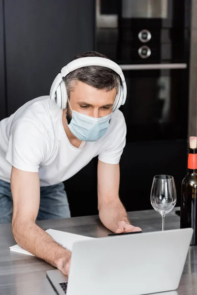 Freelancer em máscara médica e fones de ouvido usando laptop perto de copo de vinho na bancada na cozinha — Fotografia de Stock