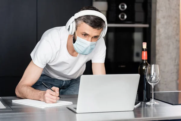 Teleworker in headphones and medical mask writing on notebook near laptop and wine on worktop in kitchen — Stock Photo