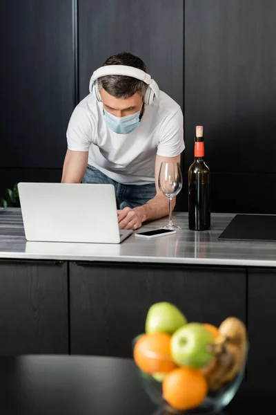 Selective focus of freelancer in headphones and medical mask using laptop near wine on kitchen worktop — Stock Photo