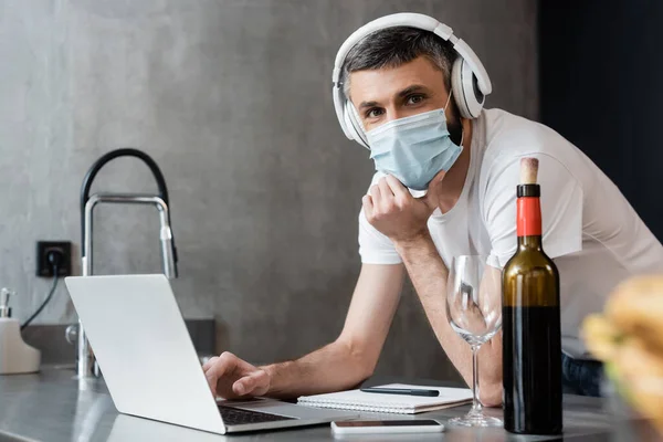 Enfoque selectivo del hombre en auriculares y máscara médica utilizando el portátil cerca del vino en la encimera de la cocina - foto de stock
