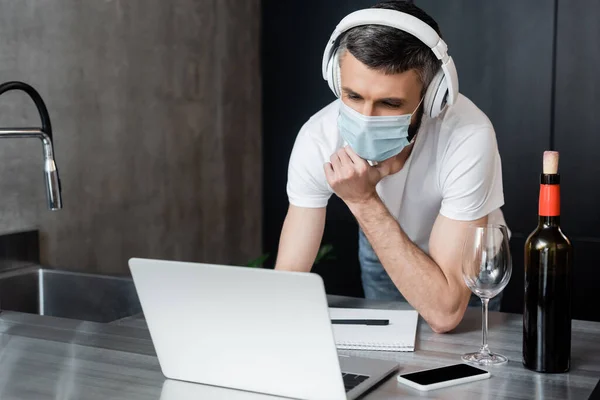 Freelancer em fones de ouvido e máscara médica olhando para laptop perto de vinho e smartphone na bancada da cozinha — Fotografia de Stock