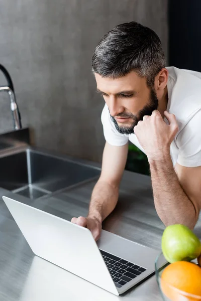 Selective focus of handsome freelancer using laptop near fruits on kitchen worktop — Stock Photo