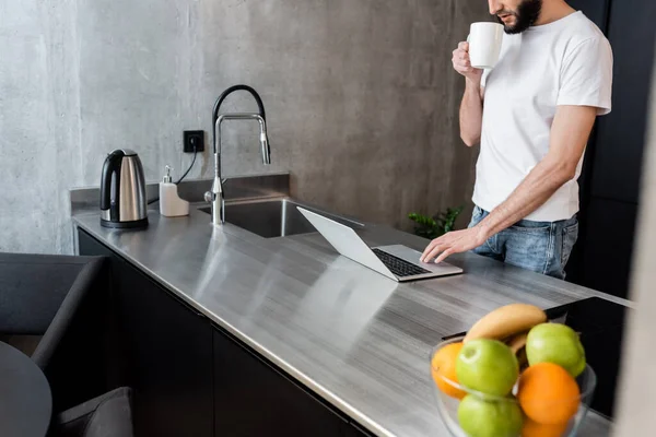 Vista recortada del freelancer sosteniendo la taza y utilizando el ordenador portátil en la encimera de la cocina - foto de stock