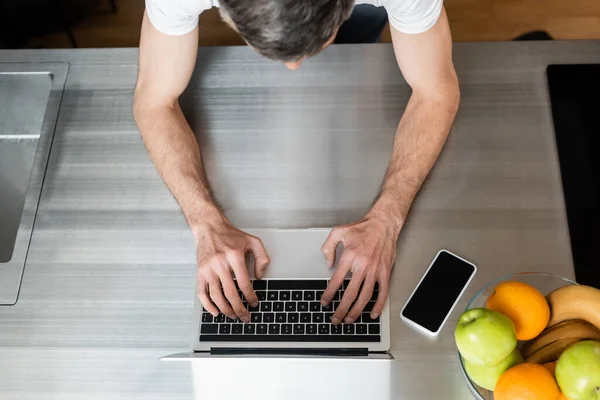 Overhead view of freelancer using laptop near smartphone and fruits on kitchen worktop — Stock Photo