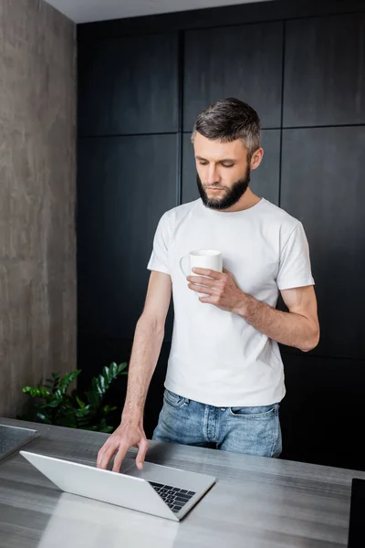 Teletrabajo guapo usando el ordenador portátil y taza de celebración en la cocina - foto de stock