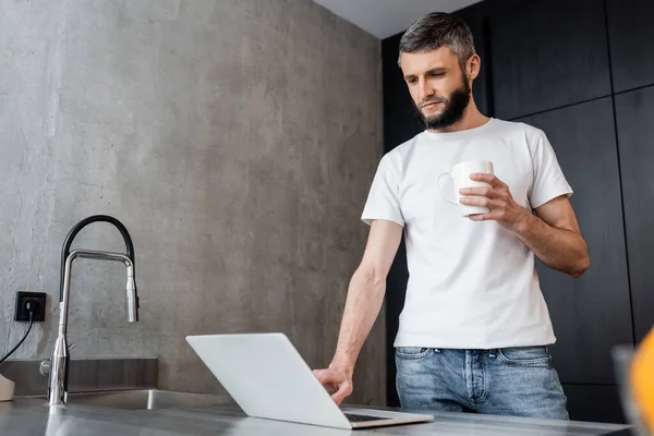 Enfoque selectivo del apuesto freelancer usando laptop y sosteniendo taza en la cocina - foto de stock