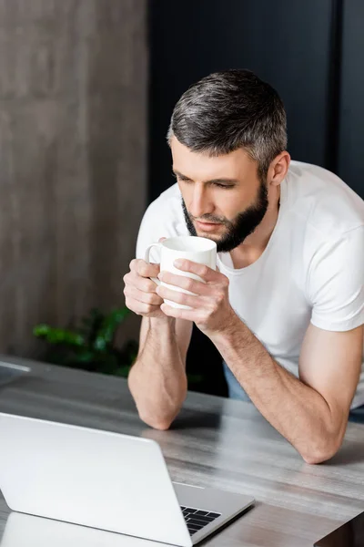 Bonito homem segurando copo e olhando para laptop na bancada da cozinha — Fotografia de Stock