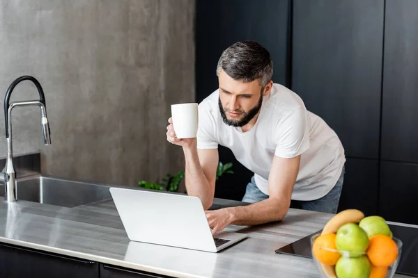 Selective focus of handsome man with cup using laptop near fruits on worktop in kitchen — Stock Photo
