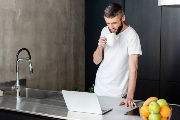 Enfoque selectivo del hombre sosteniendo taza y mirando a la computadora portátil en la cocina — Stock Photo