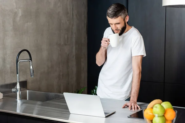 Selective focus of man holding mug and looking at laptop in kitchen — Stock Photo