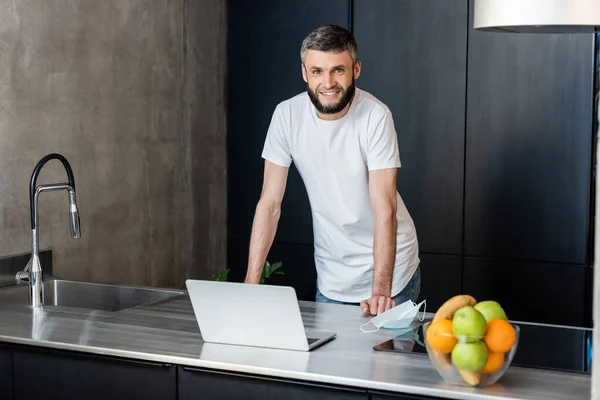 Handsome freelancer smiling at camera near laptop and medical mask on kitchen worktop — Stock Photo