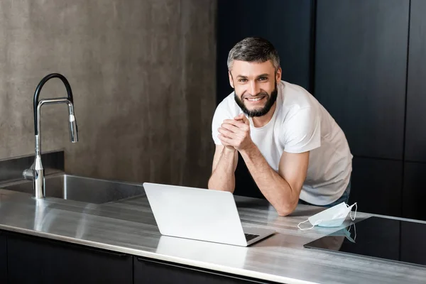 Bello freelance sorridente alla macchina fotografica vicino alla maschera medica e laptop sul piano di lavoro della cucina — Foto stock