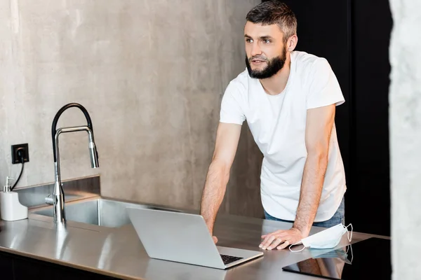 Handsome man using laptop near medical mask on worktop in kitchen — Stock Photo