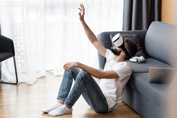 Вид сбоку на человека, использующего VR-гарнитуру рядом с ноутбуком и наушники на диване — стоковое фото