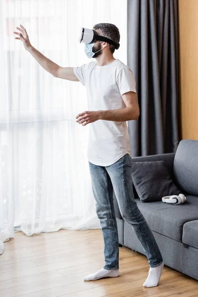 Vista laterale dell'uomo in maschera medica e auricolare vr in piedi in soggiorno — Foto stock
