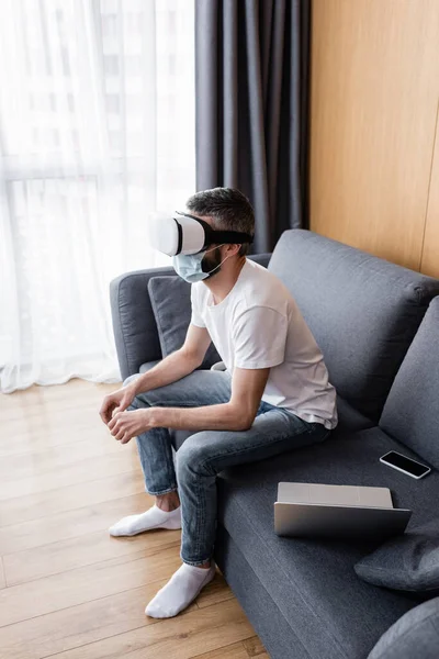 Vue latérale de l'homme en masque médical à l'aide d'un casque de réalité virtuelle près de gadgets sur le canapé — Photo de stock