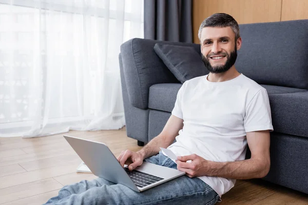 Hombre guapo sonriendo a la cámara mientras usa el ordenador portátil y la tarjeta de crédito en el suelo en casa - foto de stock