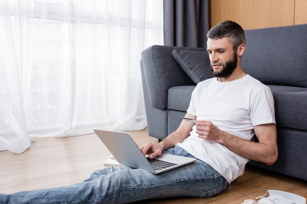 Bonito homem segurando cartão de crédito e usando laptop perto de máscara médica no chão em casa — Fotografia de Stock