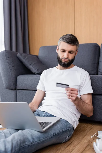 Hombre con tarjeta de crédito y portátil cerca de máscara médica y auriculares en el suelo en la sala de estar - foto de stock