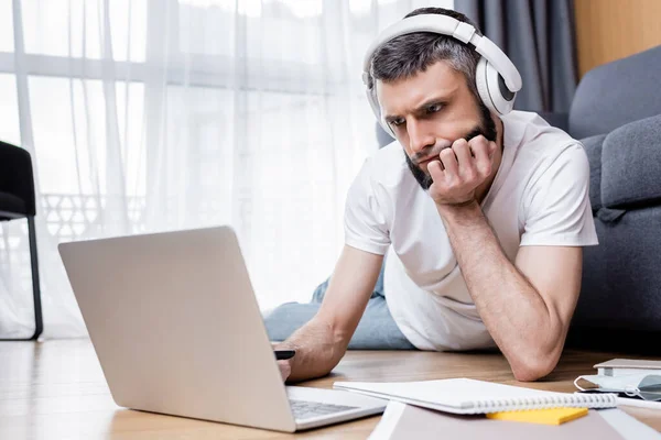 Nachdenklicher Mann mit Kopfhörer nutzt Laptop in der Nähe von Schreibwaren bei Webinar zu Hause — Stockfoto