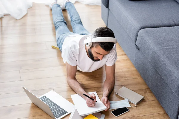 Overhead view of man in headphones writing on notebook during webinar near laptop on floor at home — Stock Photo