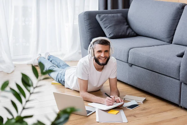 Selective focus of smiling man in headphones using stationery and laptop near medical mask on floor at home — Stock Photo