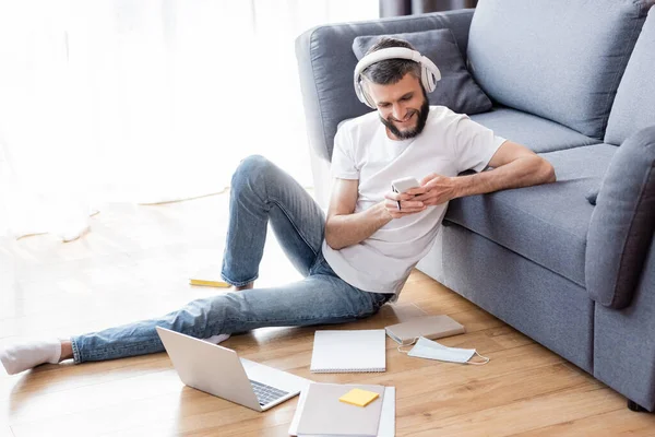 Homme souriant dans les écouteurs en utilisant un smartphone pendant le webinaire près de l'ordinateur portable et la papeterie sur le sol à la maison — Photo de stock