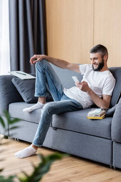 Selective focus of smiling man using smartphone near laptop, books and medical mask on couch — Stock Photo