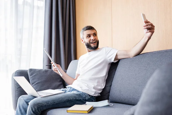 Foco seletivo de freelancer alegre tomando selfie com smartphone perto de laptop e máscara médica no sofá — Fotografia de Stock