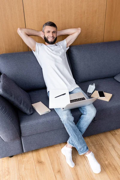 Smiling freelancer looking at camera near laptop, books and medical mask on couch — Stock Photo