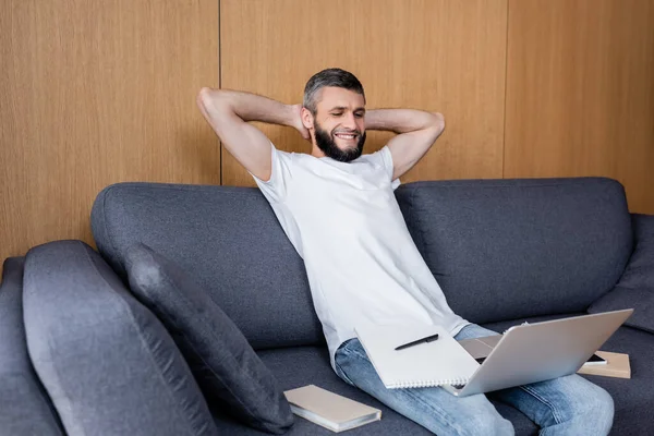 Telelavoro sorridente seduto sul divano vicino a dispositivi digitali e libri — Foto stock