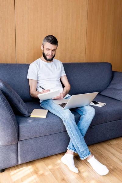 Handsome freelancer smiling while writing on notebook near digital devices and books on couch — Stock Photo