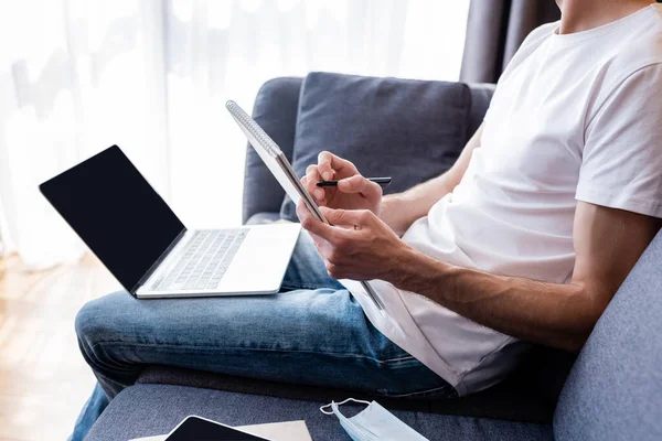 Cropped view of freelancer writing on notebook near gadgets and medical mask on couch — Stock Photo