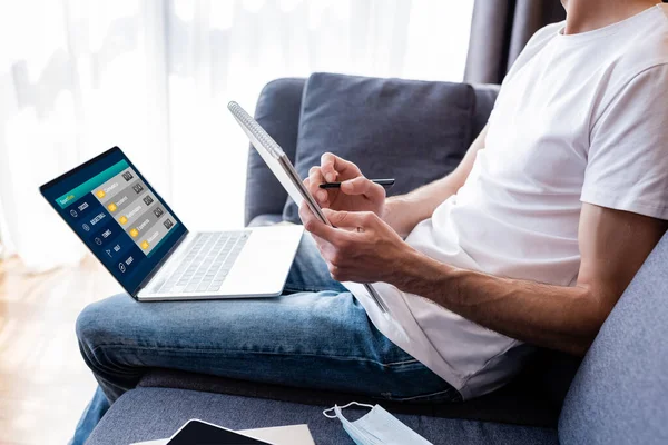 Обрізаний вид людини, що пише на ноутбуці біля ноутбука зі спортивними ставками на екрані та медичною маскою на дивані — стокове фото