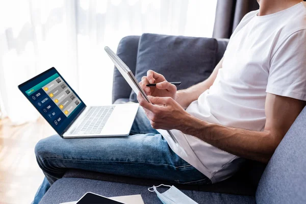 Cropped view of man writing on notebook near laptop with sport bets on screen and medical mask on couch — Stock Photo