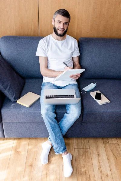 Freelancer sonriente mirando la cámara mientras escribe en un cuaderno cerca de gadgets y máscara médica en el sofá - foto de stock