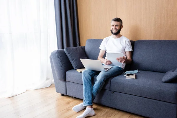 Libero professionista sorridente che tiene notebook vicino a computer portatile, libri e maschera medica sul divano a casa — Foto stock