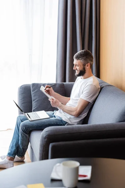 Selective focus of smiling teleworker working with laptop and notebook on sofa in living room — Stock Photo