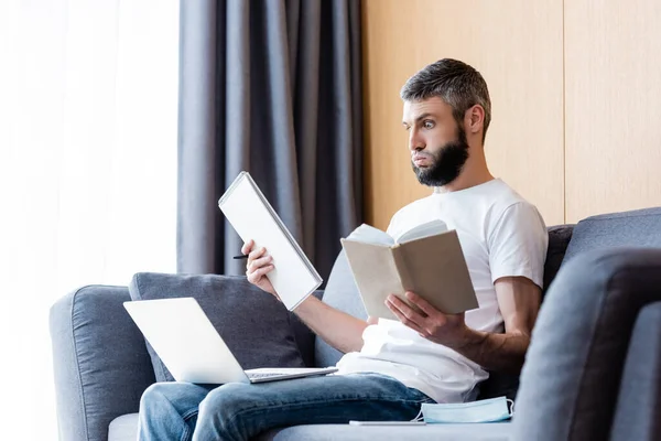 Enfoque selectivo del freelancer pensativo que sostiene el cuaderno y el libro cerca del ordenador portátil y la máscara médica en el sofá en casa - foto de stock