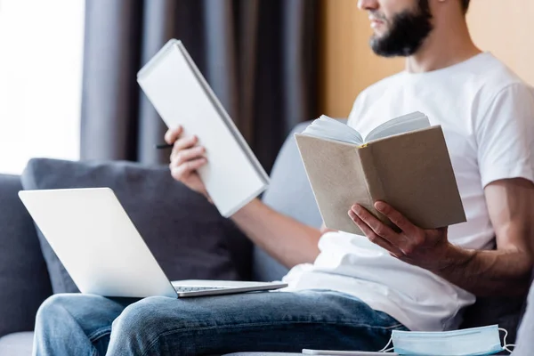 Vista recortada del freelancer que trabaja con el ordenador portátil, libro y portátil cerca de la máscara médica en la sala de estar - foto de stock