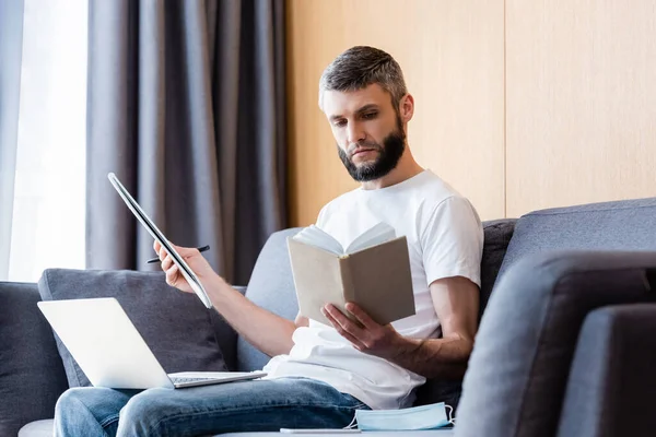 Selektiver Fokus des Telearbeiters beim Lesen von Büchern während der Arbeit mit Laptop und Notebook in der Nähe medizinischer Maske auf der Couch — Stockfoto