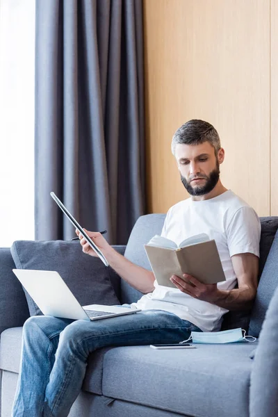 Freiberuflerin liest Buch, während sie in der Nähe digitaler Geräte und medizinischer Maske auf dem Sofa im Wohnzimmer arbeitet — Stockfoto