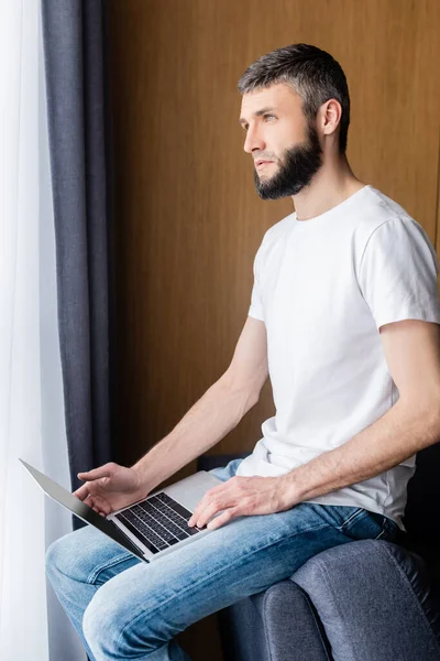 Vista lateral del freelancer mirando hacia otro lado mientras sostiene el ordenador portátil en casa - foto de stock