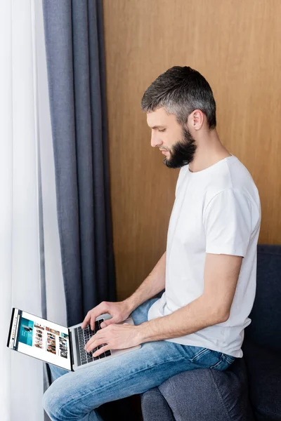 KYIV, UKRAINE - APRIL 10, 2020: Side view of man using laptop with amazon website on couch at home — стокове фото