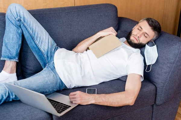 Freelancer in medical mask sleeping near book and digital devices on couch — Stock Photo