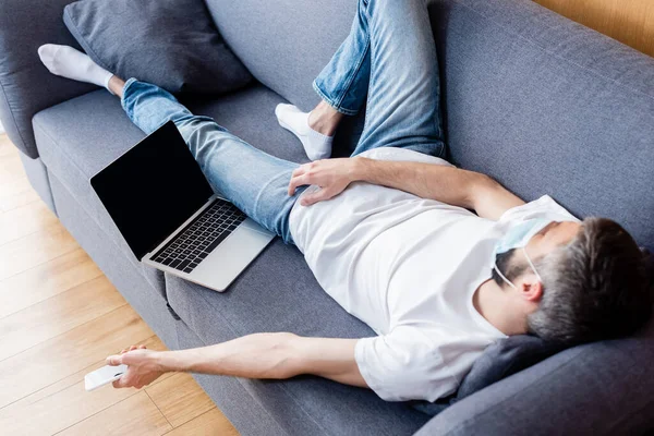 Человек в медицинской маске держит смартфон, пока спит на диване рядом с ноутбуком — стоковое фото