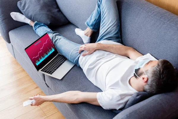 Mann mit medizinischer Maske hält Smartphone im Schlaf auf Couch neben Laptop mit Diagrammen auf dem Bildschirm — Stockfoto