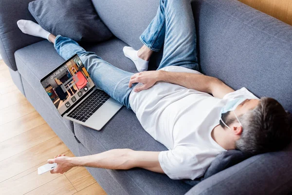 Hombre con máscara médica sosteniendo teléfono inteligente mientras duerme cerca de la computadora portátil con sitio web de reserva en línea en la pantalla - foto de stock
